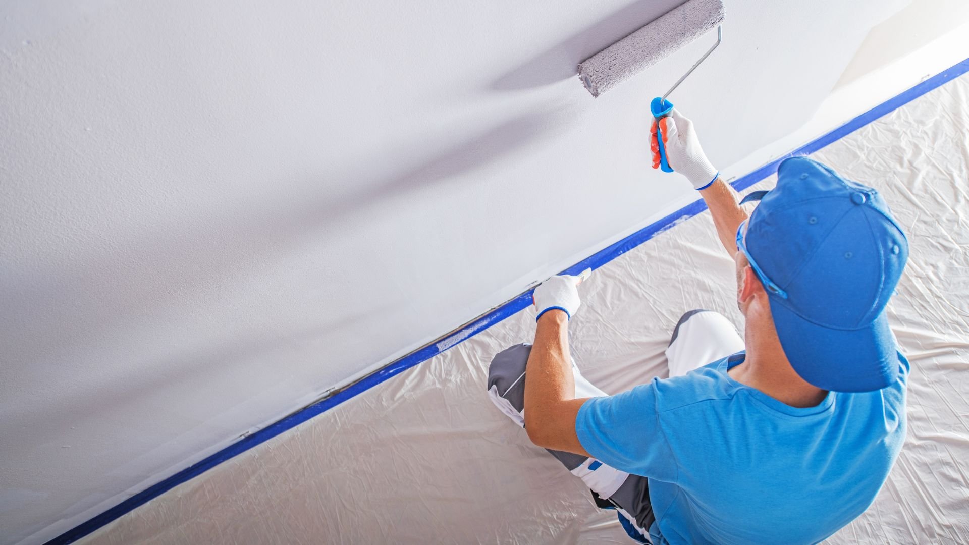 Painter in blue attire painting ceiling with roller and blue tape
