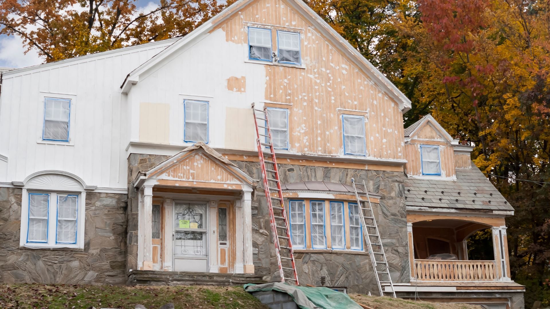 Large house under renovation with ladders, autumn trees in background