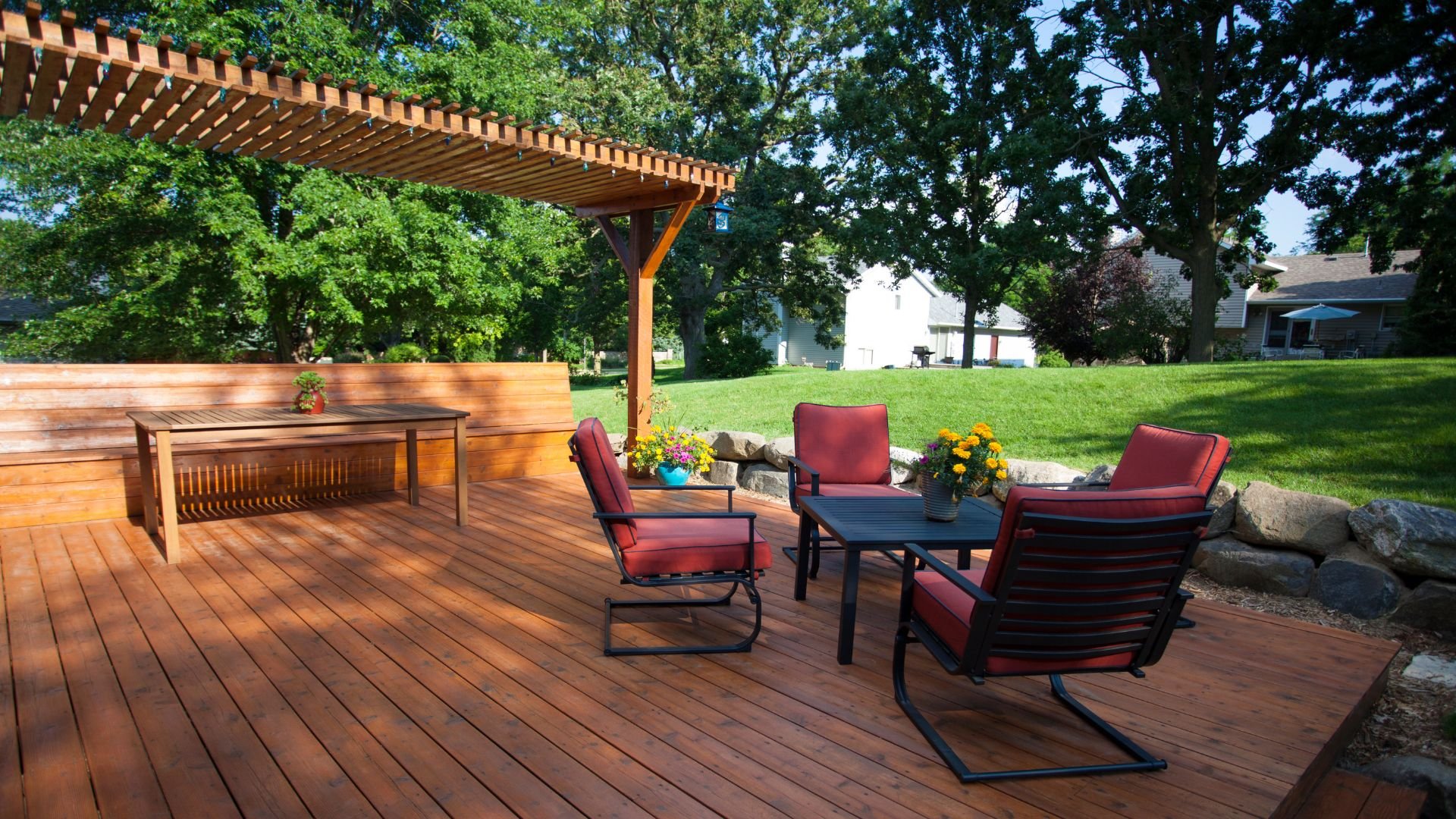 Wooden deck with red chairs, pergola, and landscaped backyard on sunny day