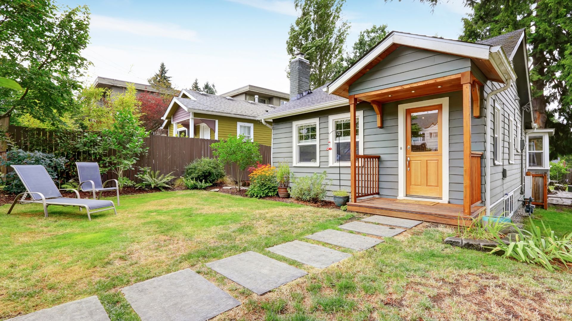 Gray house with wooden porch, lawn, and lounge chairs in residential neighborhood
